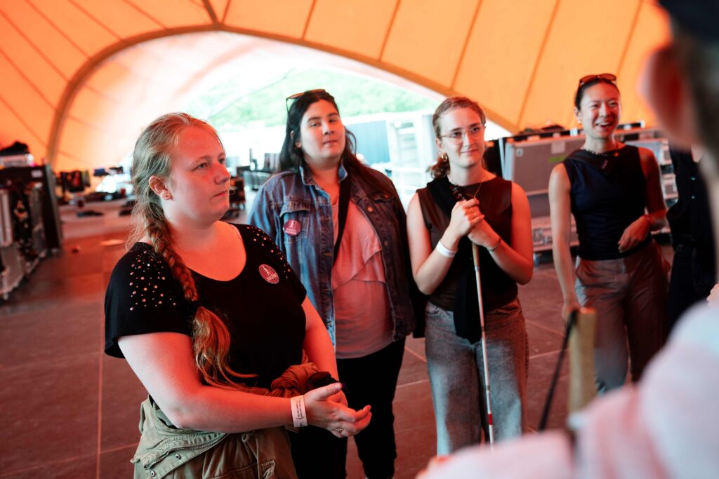 I et orange skær fra den orange scene på Roskilde festival, står tre unge kvinder og lytter til nogen fortælle.