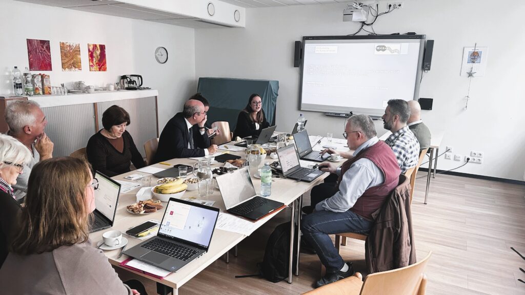 Et mødelokale, hvor projektgruppen med 8 deltagere arbejder og man ser en PowerPoint på skærm for enden af bordet.