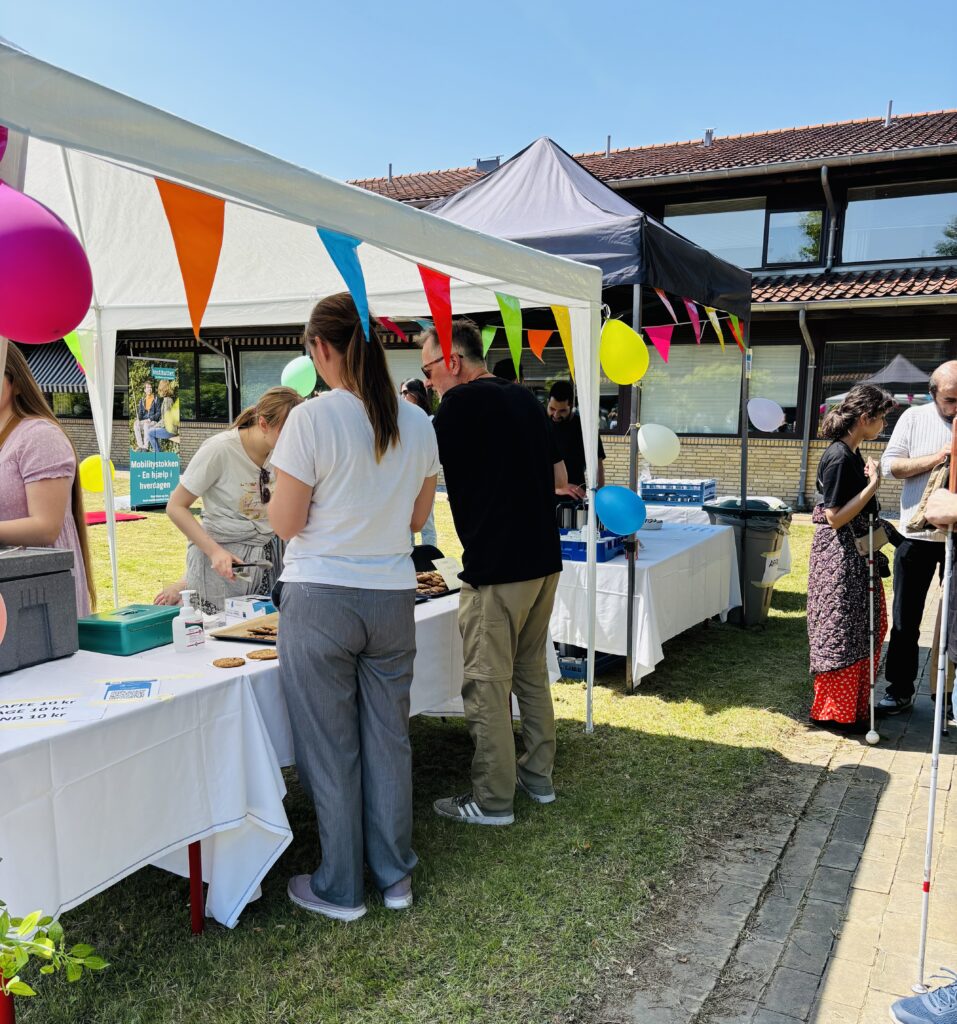 Festivalgæster står ved boden med farvede vimpler og køber hjemmebagte cookies.