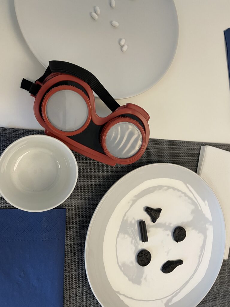 Hvidt bord dækket op med mørk dækkeserviet, hvid tallerken med sorte lakridser på og et par simulationsbriller.