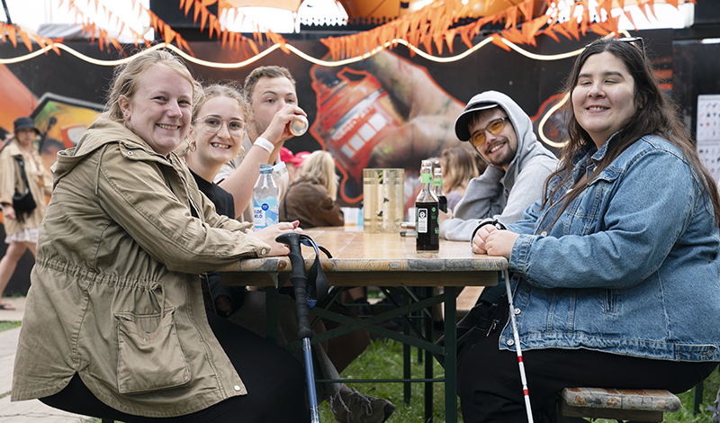 Unge fra sommertræf, på Roskilde festival. De sidder sammen ved et bord og drikker en drink med graffiti i baggrunden.